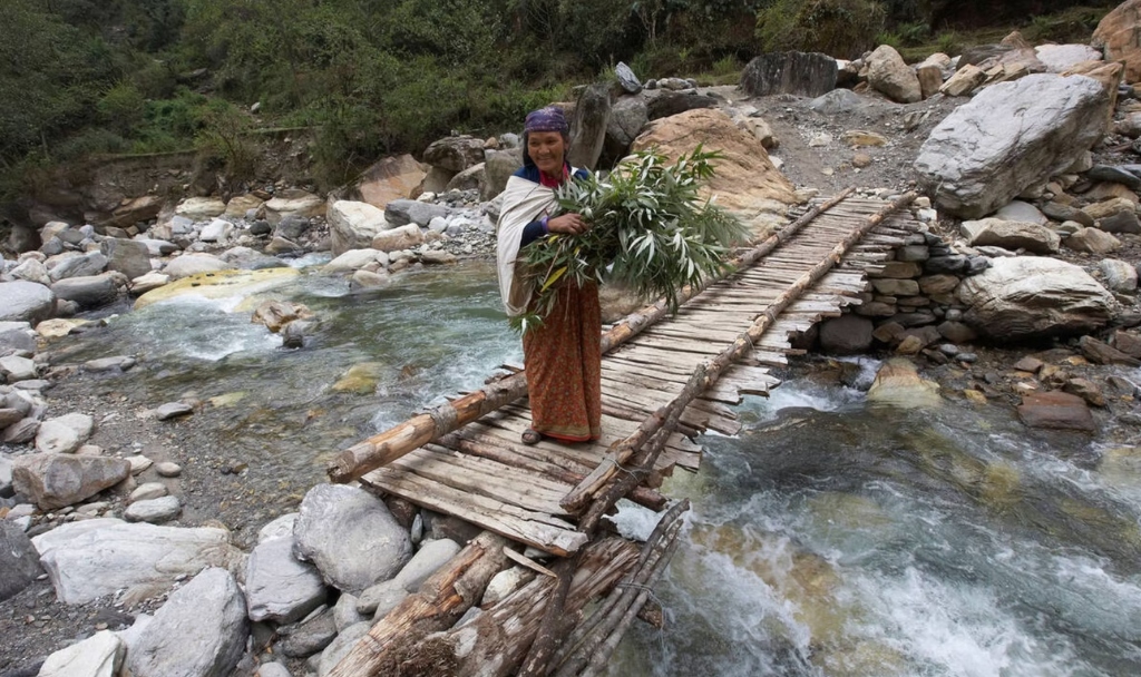 Elderly Nepali woman standing on a rustic wooden suspension bridge over a glacier river
