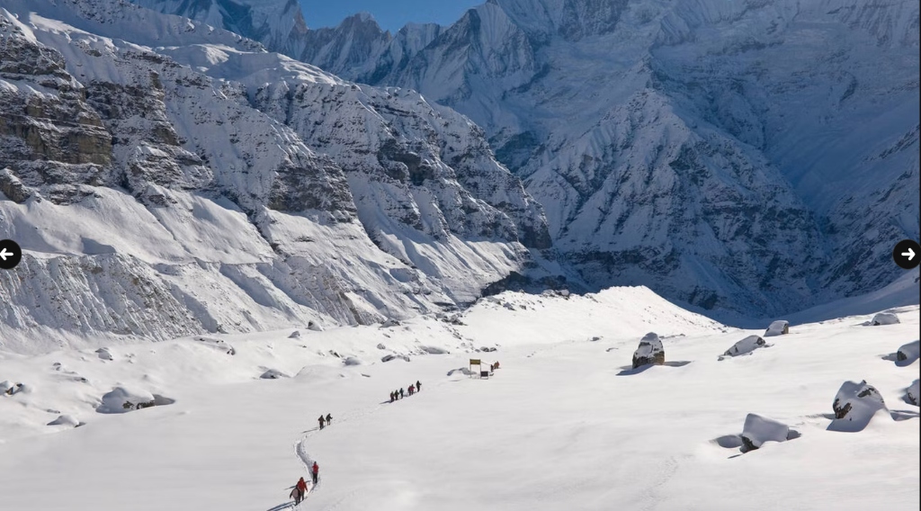 Trekkers walking in a single file line through a wide, deep snow valley between steep peaks